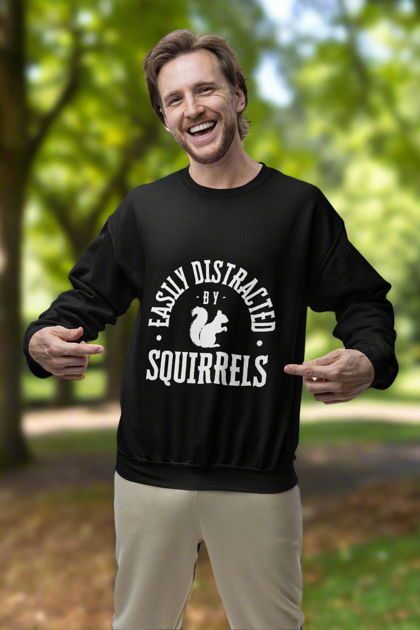A joyful man in a studio pointing at his black Gildan sweatshirt, featuring the playful text 'Easily Distracted by Squirrels' with a squirrel graphic. The sweatshirt exudes humor and cozy vibes, perfect for casual and quirky fashion lovers.