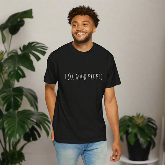 A man smiling while standing at home wearing a black 'I See Good People' unisex t-shirt. The minimalist white text design and relaxed fit highlight the shirt's casual and positive vibe.