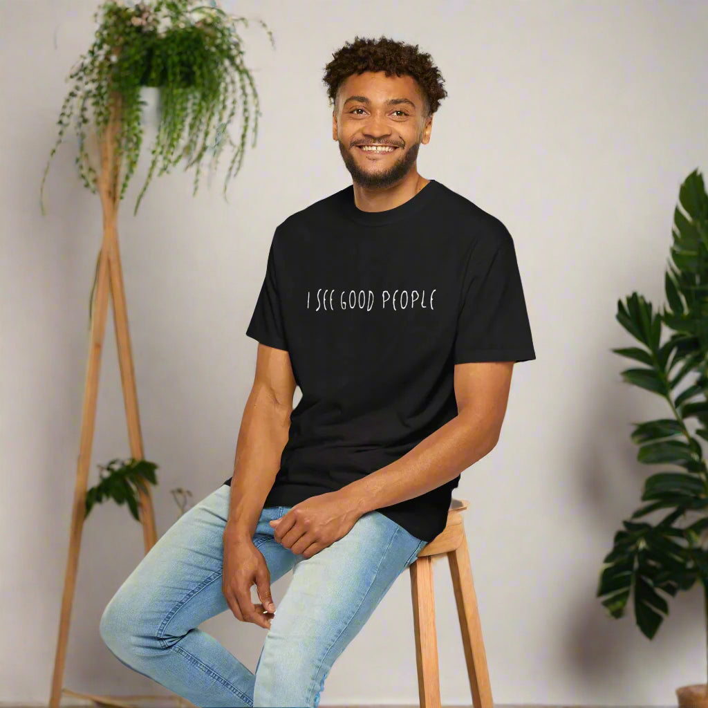 A smiling man wearing a black 'I See Good People' unisex t-shirt, seated on a wooden stool. The minimalist white text design stands out on the garment-dyed fabric, showcasing the t-shirt's relaxed fit and casual style. Perfect for spreading positivity and good vibes.