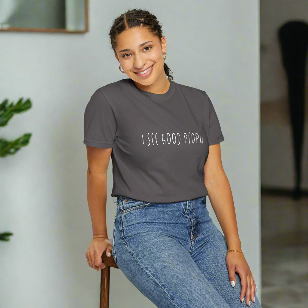 A smiling woman sitting on a stool wearing a gray 'I See Good People' unisex t-shirt. The minimalist white text design on the garment-dyed fabric emphasizes the shirt's relaxed fit and casual style, perfect for promoting positivity and good vibes.