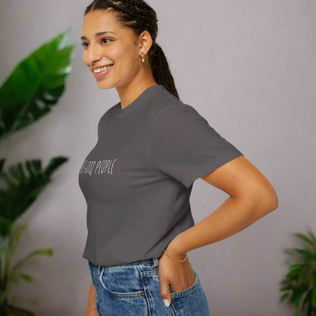 A smiling woman wearing a gray 'I See Good People' unisex t-shirt, styled casually with denim jeans. The minimalist white text design on the garment-dyed fabric highlights the shirt's relaxed fit and positive message, perfect for everyday wear.